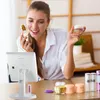 A woman is applying makeup in front of a lighted mirror, with several cosmetic products on the table. Voir les images du produit