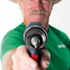 A man in a green shirt and hat holds a drill directly towards the camera. Podgląd zdjęcia produktu