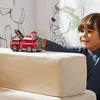 A young boy plays with a red toy fire truck on a light-colored surface, with cartoon dog drawings on the wall behind him. Produktbild-Vorschau