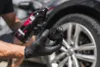 A person wearing black gloves is holding a black applicator bottle and a black applicator with a white logo, with a car wheel in the background. Anteprima dell’immagine prodotto