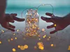 Hands reaching for a glass jar filled with fairy lights on a beach. Náhled obrázku produktu