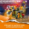 A LEGO City Stunt Park playset with a motorcycle, spider cage, and minifigure, set against a backdrop of a stadium crowd and stunt ramps. Podgląd zdjęcia produktu