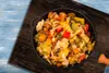 A close-up overhead view of a pan filled with a colorful vegetable stir-fry, placed on a wooden surface with a blue background. Produktbild-Vorschau