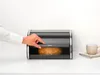 A hand is opening a black bread bin with a loaf of bread inside, on a white countertop. Podgląd zdjęcia produktu
