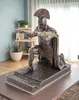 A bronze-colored statue of a Roman centurion kneeling with a spear, placed on a table in a room with a bookshelf and plants in the background. Produktbild-Vorschau