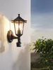 A black outdoor wall lantern is mounted on a textured white wall, with a lush green tree and a hazy landscape visible in the background under a blue sky. Náhľad obrázku produktu