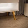 A close-up view of the lower part of a white cabinet with gold legs, resting on a wooden floor. Produktbild-Vorschau