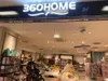 The storefront of a retail store named "360HOME" is shown. Inside the store, shelves are filled with various home goods and toys. A baby stroller is parked near the entrance. Produktbild-Vorschau