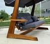A wooden swing set with dark gray cushions is shown outdoors on a paved surface, with a building and greenery in the background. Podgląd zdjęcia produktu