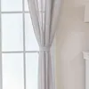 A close-up of a light gray curtain tied back with a fabric band, hanging in front of a window with white mullions. Produktbild-Vorschau