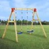 A wooden swing set with two blue swings is set up in a grassy field with trees in the background and a blue sky above. Podgląd zdjęcia produktu