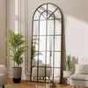 A full-length arched mirror with a black metal frame is placed in a living room setting, reflecting furniture and curtains. A potted palm and a snake plant are visible. Produktbild-Vorschau