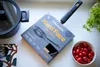 A Fiskars frying pan in its packaging is displayed on a white surface, next to a pot, a patterned cloth, and a bowl of cherry tomatoes. Podgląd zdjęcia produktu