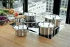 A set of five stainless steel pots and pans are arranged on a modern induction stovetop, with fresh vegetables and ingredients visible on a wooden countertop in the background. Produktbild-Vorschau