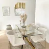 A modern dining room with a glass-top dining table, gold chandelier, and beige chairs. Produktbild-Vorschau