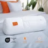 A white bolster pillow is placed on a bed with white and orange pillows in the background. Produktbild-Vorschau