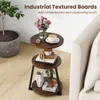 A round, three-tiered side table with a rustic wood finish and black metal legs, placed next to a sofa and a potted plant. It is decorated with a vase of flowers, a cup of coffee, books, and decorative items. Podgląd zdjęcia produktu