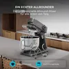 A kitchen mixer with its accessories is displayed on a countertop, with kitchen cabinets and a stove in the background. Text overlays highlight features. Produktbild-Vorschau