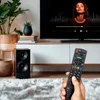 A hand holding a remote control in front of a TV and speaker setup. Voir les images du produit