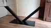 A black V-frame table base is shown on a wooden floor in front of a white wall and a wooden door. Produktbild-Vorschau