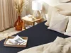A bedroom scene with a dark blue fitted sheet on a bed, decorated with beige pillows and a woven tray with magazines. Produktbild-Vorschau