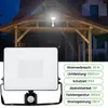 A floodlight with a motion sensor is shown in the foreground, with a wooden carport and a house in the background. Náhled obrázku produktu