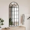 A tall, arched mirror with a black metal frame, resembling a window, is mounted on a light-colored wall above a wooden dresser. A plant is visible to the left, and a scarf and hanger are on the wall to the right. Anteprima dell’immagine prodotto