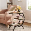A narrow, C-shaped side table with three shelves is placed next to a pink sofa. The table holds a vase of yellow flowers, a glass of amber liquid, a woven basket, a decorative shell, and a book. Voir les images du produit