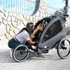 A woman is interacting with two children in a black bicycle trailer. Produktbild-Vorschau