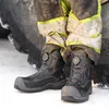 Close-up of a person wearing work boots and protective pants, standing in snow next to a large tire. Produktbild-Vorschau