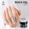 A close-up of a hand with long, light pink acrylic nails, wearing a silver ring, with a jar of Victoria Vynn Build Gel in the foreground. Náhled obrázku produktu