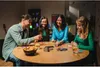 Four people are sitting around a wooden table playing a card game. Anteprima dell’immagine prodotto