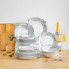 A collection of white ceramic dinnerware with a geometric gold and gray pattern, arranged on a wooden surface with a cutting board, a small vase with berries, and a bottle of champagne with a glass. Produktbild-Vorschau