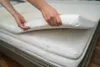Hands are shown adjusting a white mattress topper on top of a mattress. Náhľad obrázku produktu