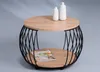 A round coffee table with a wood top and a black metal wire base, featuring two shelves. A magazine and two decorative vases are placed on the table. Produktbild-Vorschau