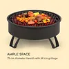 A black metal fire pit filled with grilled food, including skewers, corn on the cob, and steaks, resting on four black legs. The background is a plain light yellow. Produktbild-Vorschau
