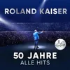 A man in a blue suit stands on a stage in front of a large audience, illuminated by spotlights. Text indicates it's a live album celebrating 50 years of hits. Voir les images du produit