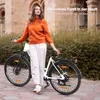 A woman stands next to a white e-bike on a paved path with a building and bushes in the background. Produktbild-Vorschau