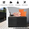 A black rattan storage box with orange cushions and a blanket on top, placed on a white deck. The background shows a white wall and a dark fence. Voir les images du produit