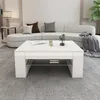 A white coffee table is placed in a living room setting with a curved sofa and a plant. Náhled obrázku produktu