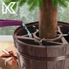 Watering a Christmas tree in a brown tree stand, with a gift box nearby. Voir les images du produit