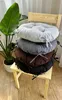 A stack of four round seat cushions in different colors (light gray, gray, brown, and black) are placed on a wooden chair next to a large potted plant. Produktbild-Vorschau