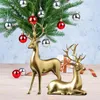 Two golden reindeer figures are placed on a wooden surface in front of a decorated Christmas tree with red and silver ornaments. Produktbild-Vorschau