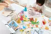 A child is crafting on a table with various craft supplies, including paper, felt cutouts, and party hats. An adult's hands are visible in the background, using an iron. Podgląd zdjęcia produktu