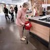 An elderly woman with a cane and a shopping basket is at a checkout counter in a store. Produktbild-Vorschau