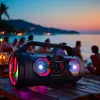 A portable Bluetooth speaker with colorful lights is placed on a wooden table on a beach at sunset, with people in the background. Produktbild-Vorschau