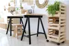 A home office or craft space with a wooden desk supported by black trestle legs, a small wooden stool, and a tall wooden crate shelf unit with a plant on top. Podgląd zdjęcia produktu