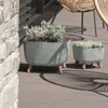 Two planters with plants on a wooden deck, with a stone wall and chair in the background. Náhled obrázku produktu