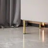 A close-up view of the bottom part of a white cabinet with gold legs, placed on a gray tiled floor next to a gray curtain. Produktbild-Vorschau