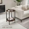 A round, two-tiered side table with a wood-look top and black metal legs is placed next to a cream-colored sofa in a living room setting. Decorative items like a plant, a carafe of water, and a book are on the table. Náhled obrázku produktu